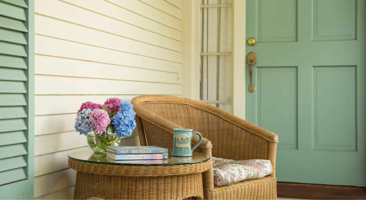 Light brown wicker chair with light paisley cushion next to light brown wicker coffee table with glass top with glass vase, pink and blue flowers, coffe mug, and books sitting on top