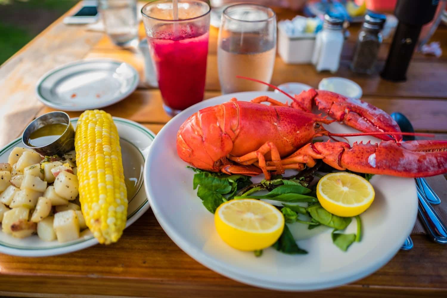 bright red lobster on a plate with greens and lemon beside an ear of yellow corn on the cob