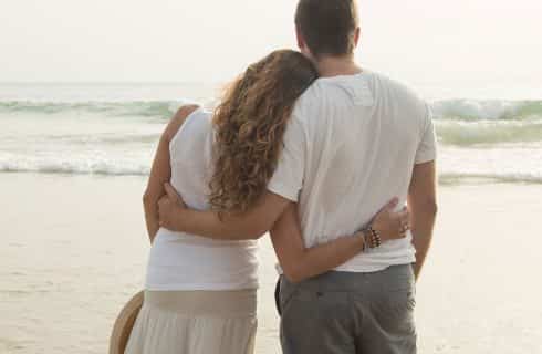 Man and woman with arms around each other standing in water on beach looking at the waves