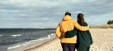 Man and woman with arms around each other wearing coats walking along the beach