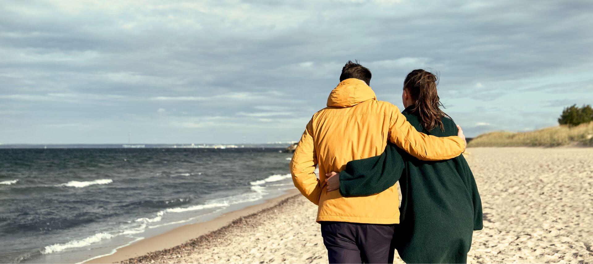 Man and woman with arms around each other wearing coats walking along the beach