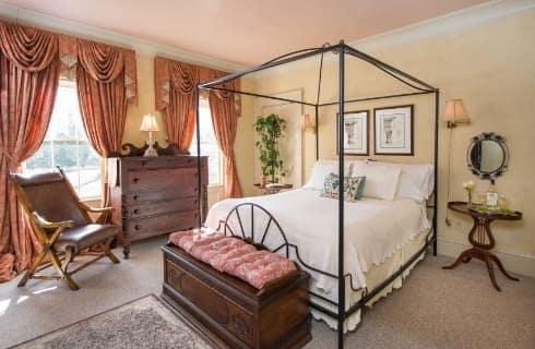 Bedroom with dark wooden furniture, rod iron bed with white bedding, wooden nightstands, yellow walls, light colored carpeting, and salmon colored drapery