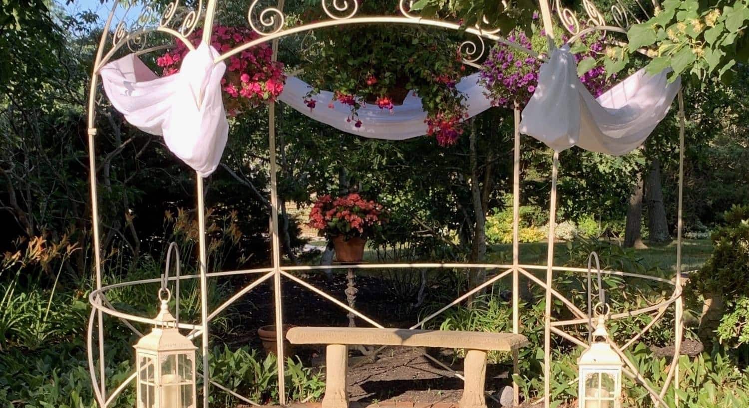 Cream rod iron gazebo decorated with hanging pink and purple flowers and draped white fabric all surrounded by green trees and shrubs with a concrete bench in the middle