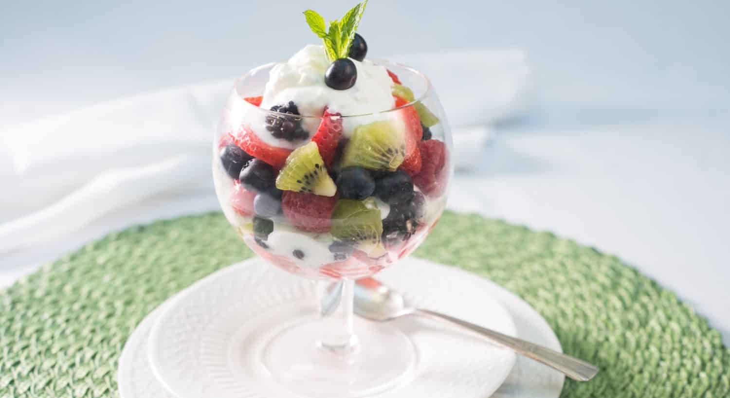 Glass fruit cup sitting on white porcelain plate filled with layers of yogurt, kiwi, strawberries, blackberries, and blueberries