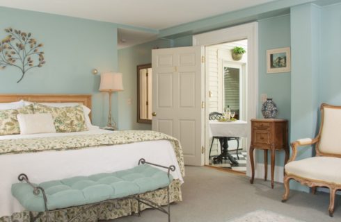 Bedroom with light mint walls and light colored carpeting, light wooden headboard with white and floral linens, rod iron bench with mint colored cushion, and light wooden nighttables with lamps