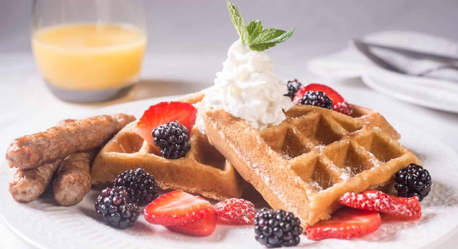 Belgian waffles topped with whipped cream, powdered sugar, strawberries, and blackberries with a side of sausage links