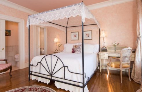 Bedroom with rod iron four-post canopy bed with white bedding, light colored wooden desk and chair in the corner, hardwood floors, peach floral wallpaper with peach drapery
