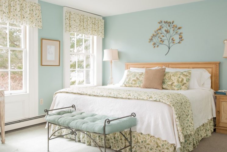 Bedroom with light mint walls and light colored carpeting, light wooden headboard with white and floral linens, rod iron bench with mint colored cushion, and light wooden nighttables with lamps