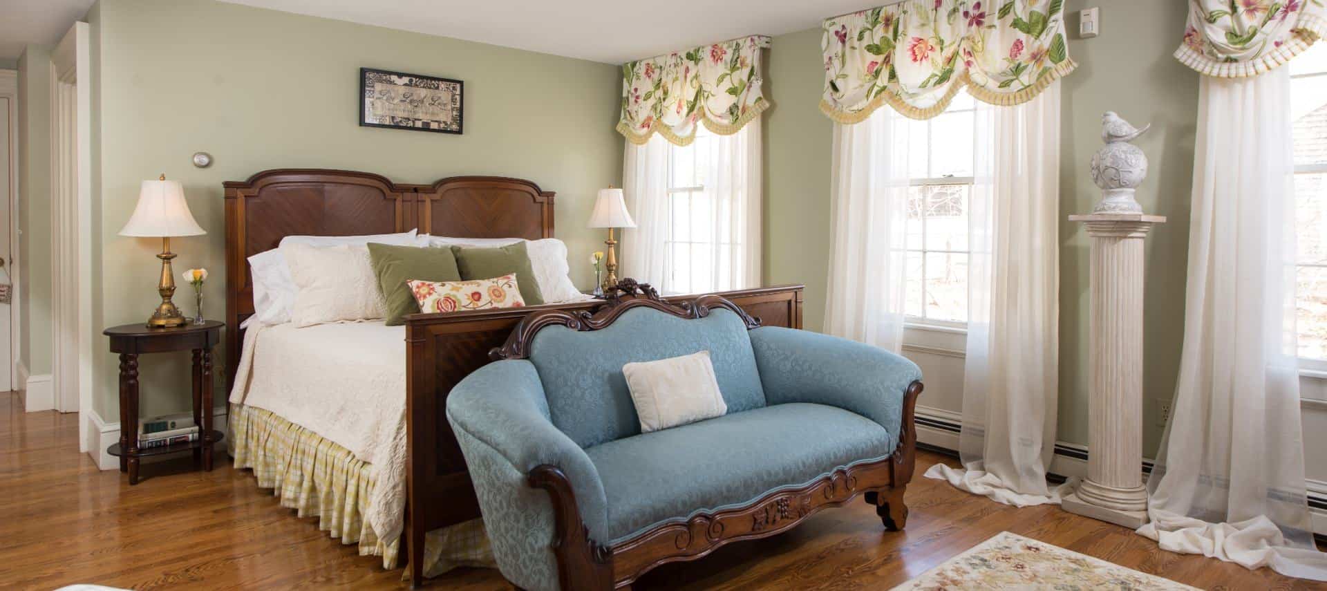 Bedroom with dark wooden furniture, cream and yellow bedding, ornate wooden love seat with blue upholstery, and sheer curtains