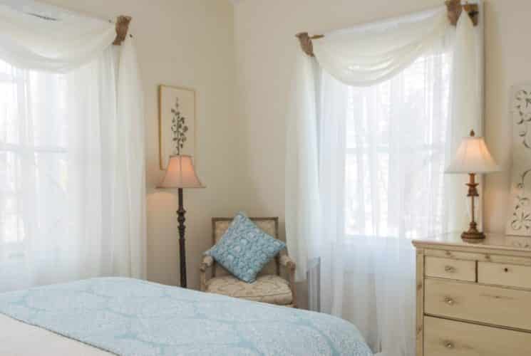 Bedroom with white and light blue bedding, upholstered vintage chair, and light wooden antique dresser with lamp, mirror, and vase full of flowers on top
