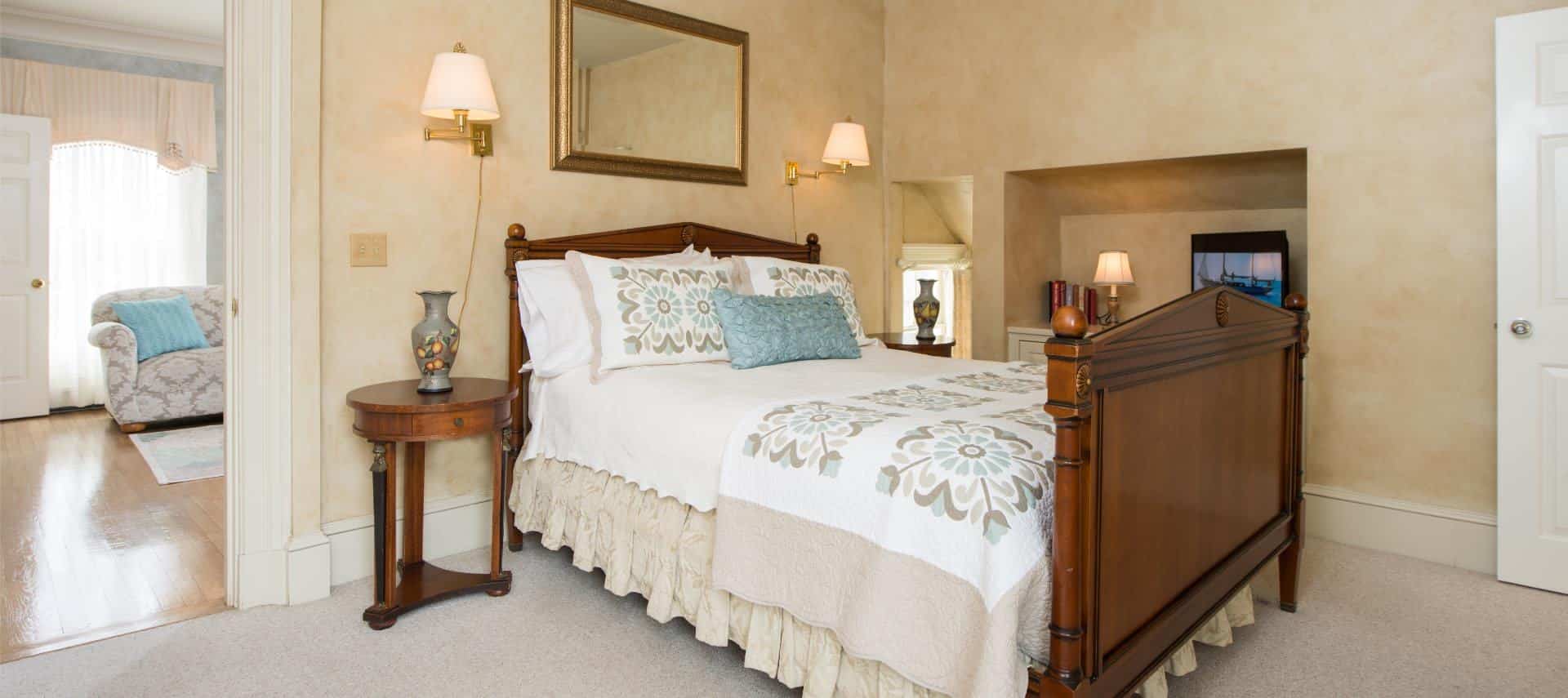 Bedroom with cream colored walls and light colored carpeting, dark wooden furntiture, white, tan, and light blue bedding, and sconce lamps attached to the wall