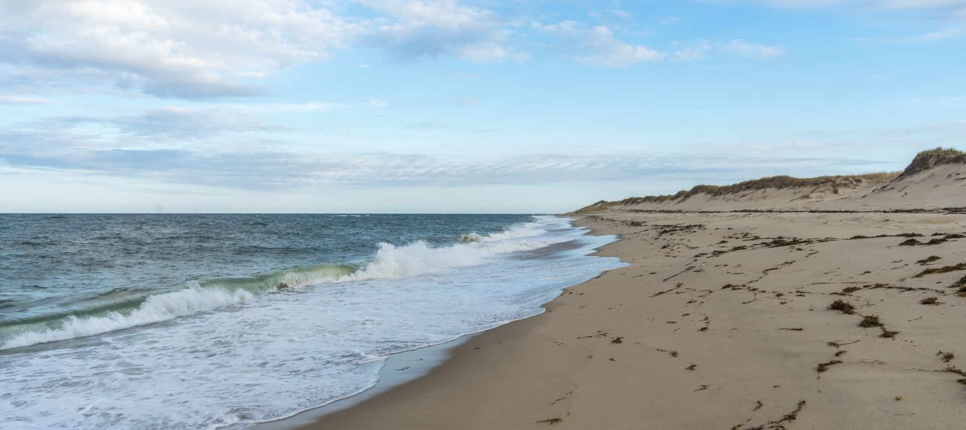 View of a sandy beach with dunes next to the ocean with gently rolling waves crashing on the beach
