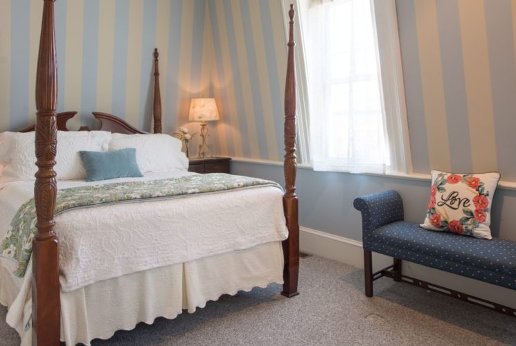 Bedroom with ornate dark wood four post bed with white bedding, wooden nightstands with iron lamps, blue and tan striped wallpaper, and light gray carpeting