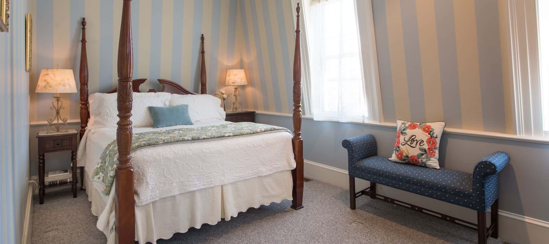Bedroom with ornate dark wood four post bed with white bedding, wooden nightstands with iron lamps, blue and tan striped wallpaper, and light gray carpeting
