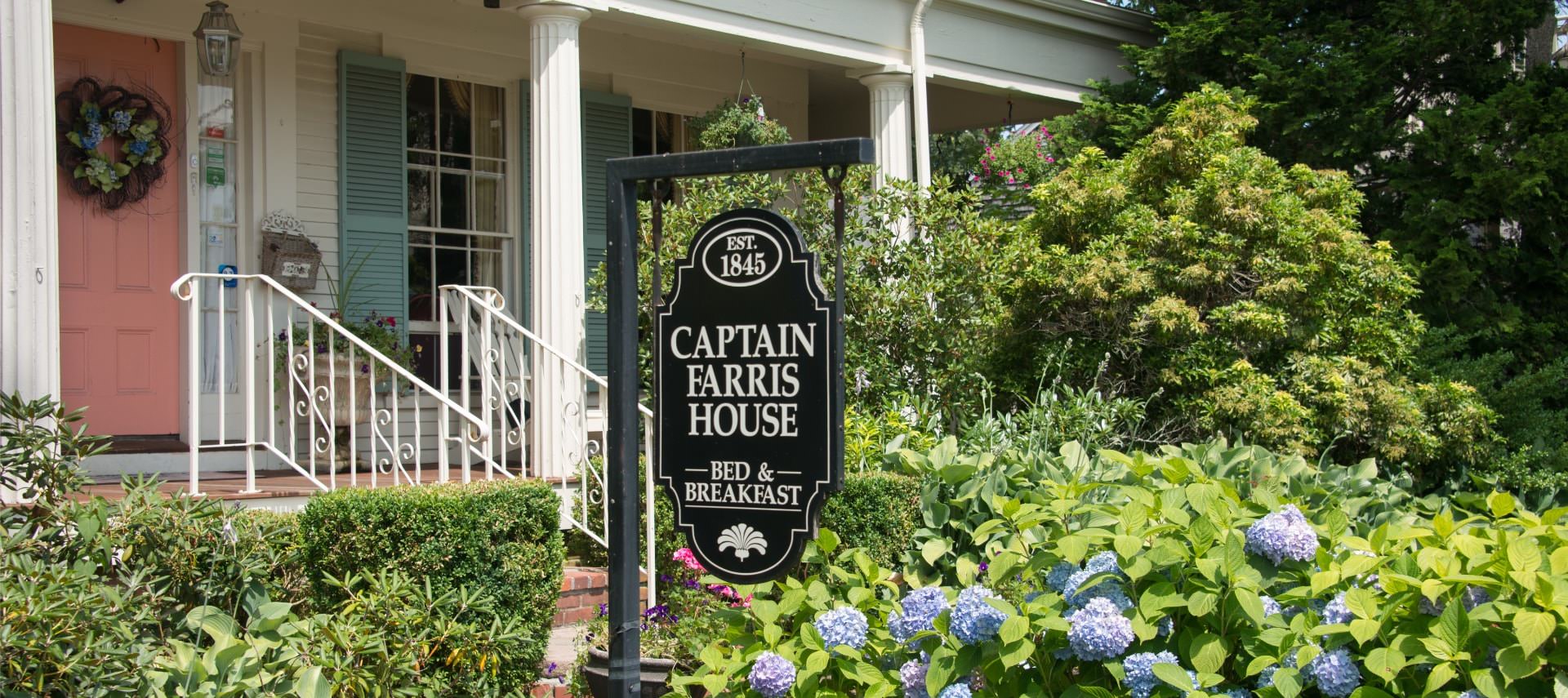 Front view of property with salmon-colored door and light turquoise shutters surrounded by shrubs, bushes, and flowers with a sign for Captain Farris House