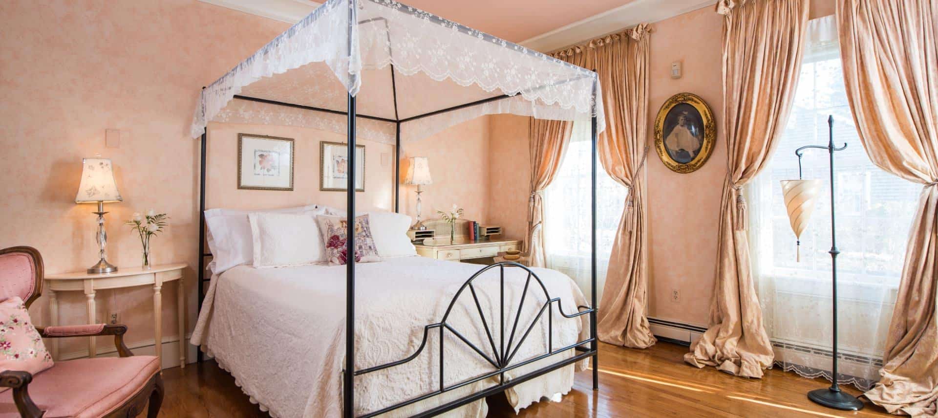 Bedroom with rod iron four-post canopy bed with white bedding, light colored wooden desk and chair in the corner, hardwood floors, peach floral wallpaper with peach drapery