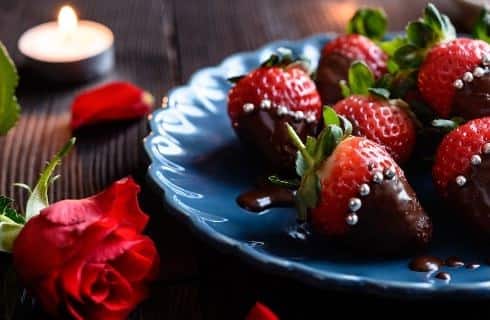 Blue porcelain plate with chocolate-covered strawberries with silver pearls on a wooden table next to a red rose and lighted tea light