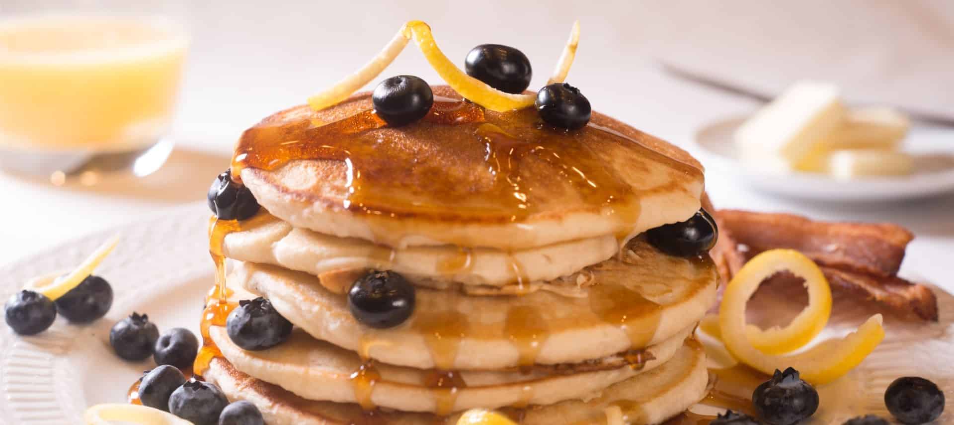 Tall stack of pancakes with syrup and blueberries on top and a side of bacon on a white porcelain plate