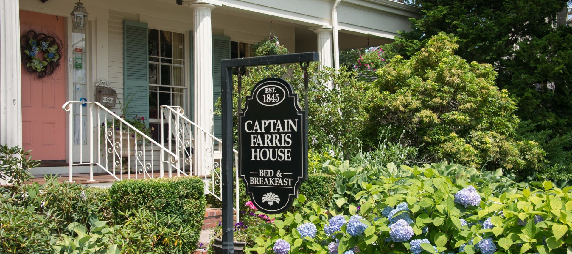 Front view of property with salmon-colored door and light turquoise shutters surrounded by shrubs, bushes, and flowers with a sign for Captain Farris House