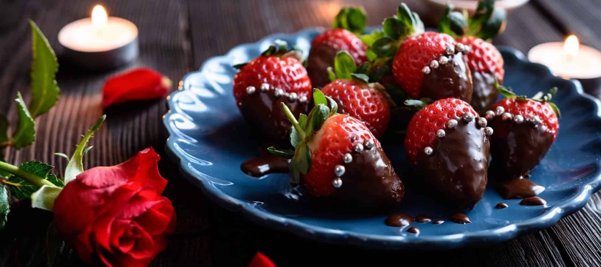 Blue porcelain plate with chocolate-covered strawberries with silver pearls on a wooden table next to a red rose and lighted tea light