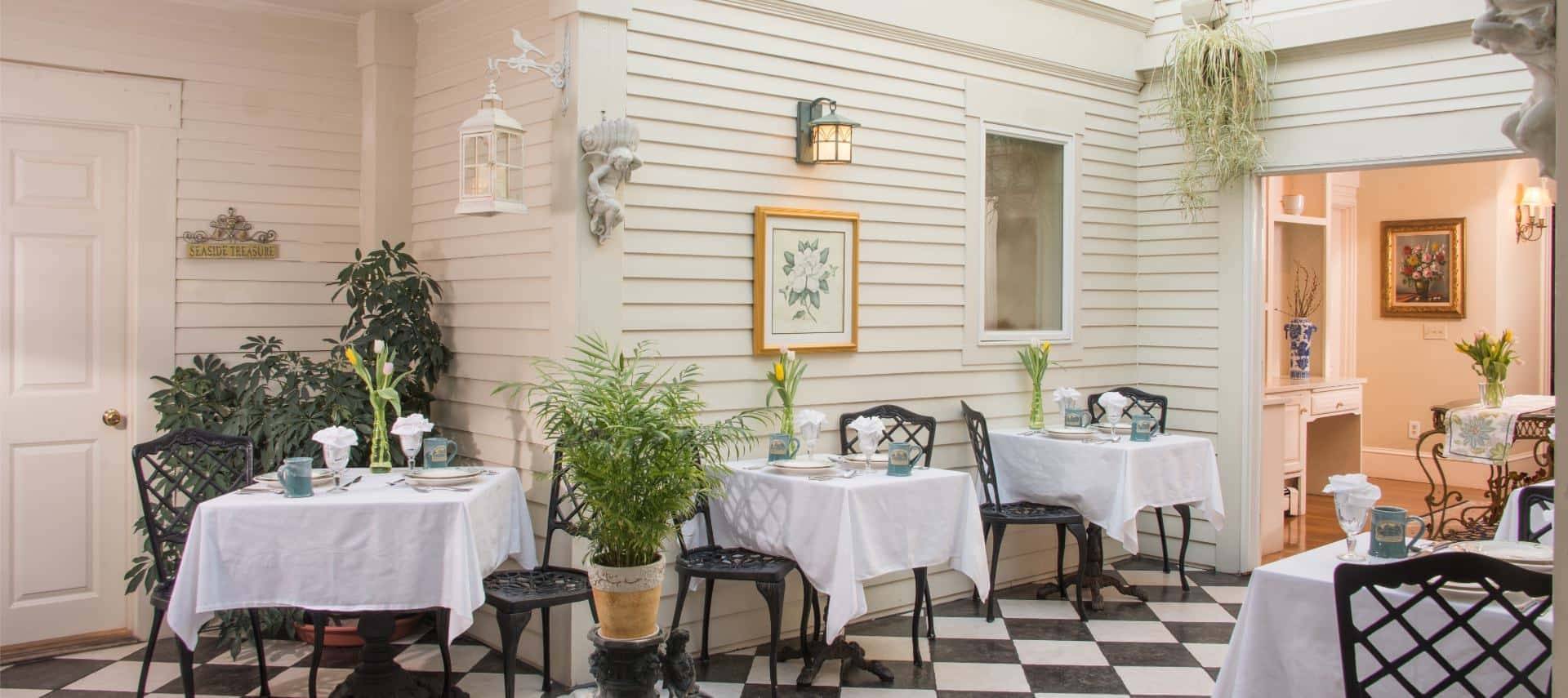 Dining area with rod iron chairs and tables with white tablecloths with white walls and black and white tiled flooring