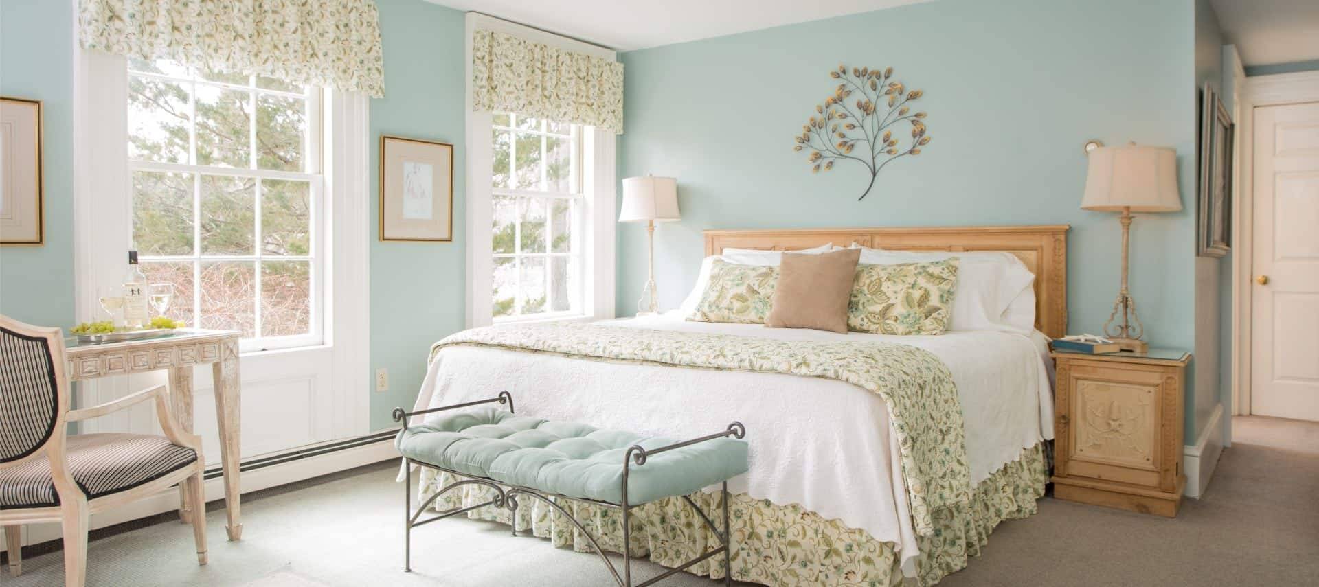Bedroom with light mint walls and light colored carpeting, light wooden headboard with white and floral linens, rod iron bench with mint colored cushion, and light wooden nighttables with lamps